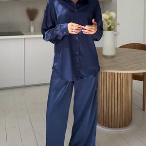 May include: A navy blue satin pajama set. The set includes a button-down shirt and matching wide-leg pants. The outfit is worn in a kitchen setting with a wooden table and white flowers in a vase.