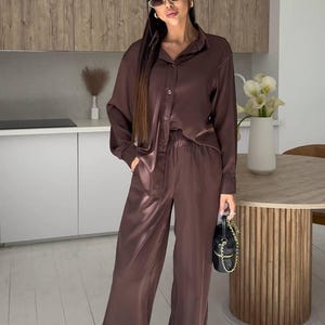 May include: A brown satin pajama set, including a button-down shirt and wide-leg pants. The outfit is worn with sunglasses and a small black purse with a gold chain. The setting is a modern kitchen.