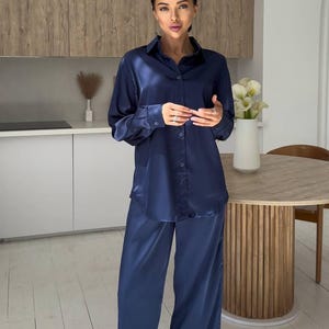 May include: A navy blue satin pajama set. The set includes a button-down shirt and matching wide-leg pants. The outfit is worn in a kitchen setting with a wooden table and white flowers in a vase.