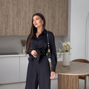 May include: A black satin two-piece outfit, consisting of a long-sleeved button-down shirt tied at the waist and wide-leg pants. The outfit is paired with a small black shoulder bag. The setting is a modern kitchen with wooden cabinets.