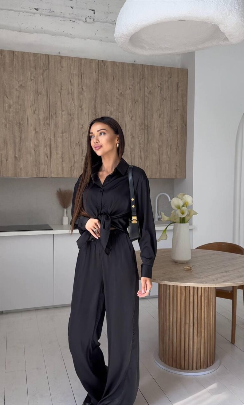 May include: A black satin two-piece outfit, consisting of a long-sleeved button-down shirt tied at the waist and wide-leg pants. The outfit is paired with a small black shoulder bag. The setting is a modern kitchen with wooden cabinets.