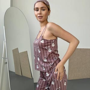 K&ouml;nnte beinhalten: Mauvefarbenes Pyjama-Set mit Hasenmuster. Das Set beinhaltet ein Tanktop, eine Hose und ein passendes Stirnband. Der Pyjama hat vertikale Streifen und wei&szlig;e Hasengesichter. Das Tanktop hat d&uuml;nne Tr&auml;ger.