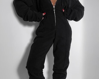 May include: A black jumpsuit with a hooded top and a full-length zipper. The jumpsuit has long sleeves and a relaxed fit. The model is wearing white socks and sneakers. The jumpsuit is made of a soft, comfortable material.