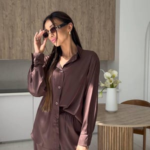 May include: A brown satin pajama set, consisting of a button-down shirt and wide-leg pants. The outfit is paired with a black quilted handbag with a gold zipper. The person is wearing sunglasses.
