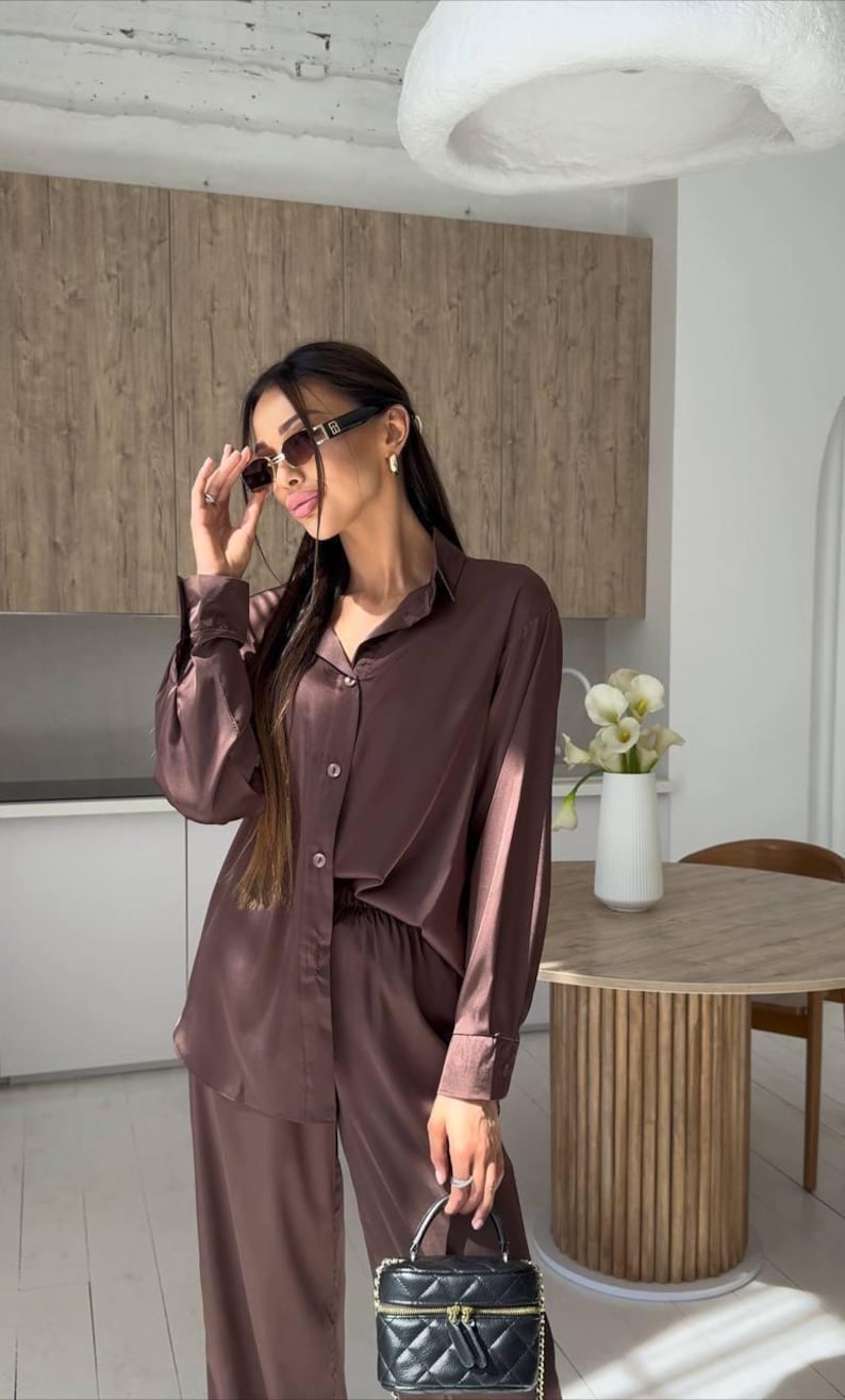 May include: A brown satin pajama set, consisting of a button-down shirt and wide-leg pants. The outfit is paired with a black quilted handbag with a gold zipper. The person is wearing sunglasses.