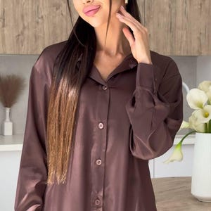 May include: A brown satin button-down shirt with long sleeves. The shirt has a classic collar and is buttoned up. The fabric has a slight sheen. The shirt is part of a matching set.