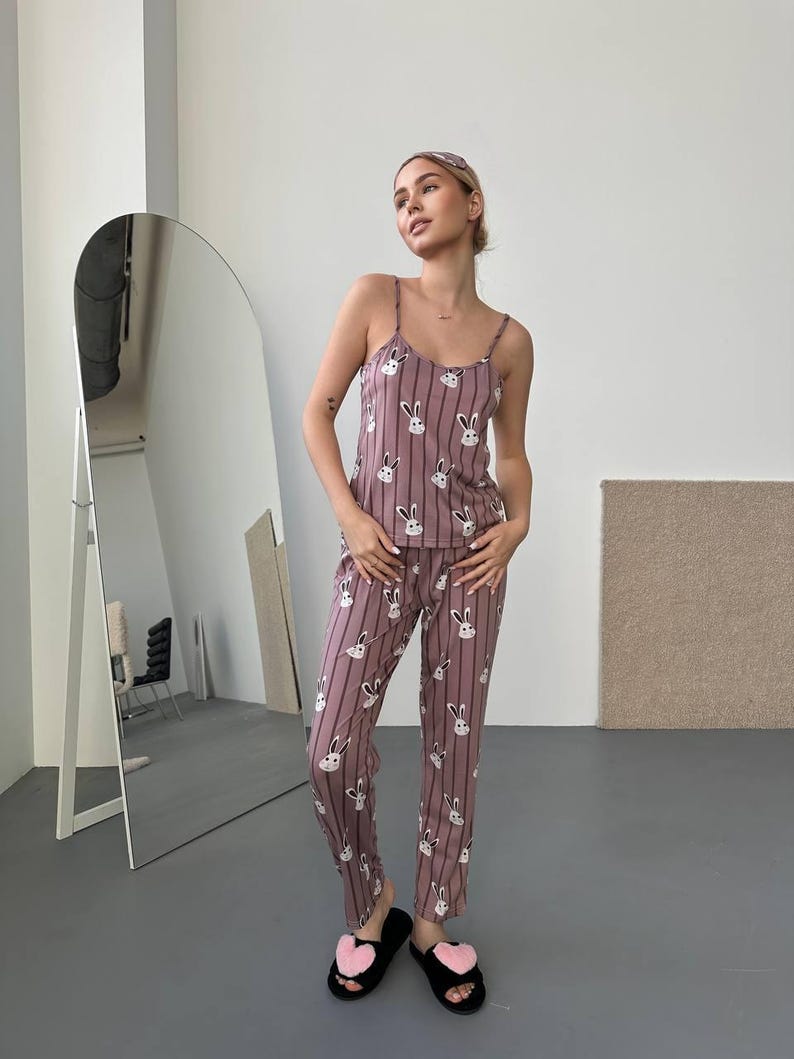 K&ouml;nnte beinhalten: Mauvefarbenes Pyjama-Set mit vertikalen Streifen und wei&szlig;en Hasengesichtern. Das Set beinhaltet ein Tr&auml;gershirt, eine Hose und ein passendes Stirnband. Die Person tr&auml;gt schwarze Hausschuhe mit rosa Herzakzenten. Der Pyjama ist aus leichtem Material.