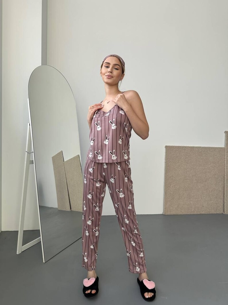 K&ouml;nnte beinhalten: Mauvefarbenes Pyjama-Set mit Hasenmuster, bestehend aus einem Tanktop und einer Hose. Das Set hat vertikale Streifen und wei&szlig;e Hasengesichter. Das Model tr&auml;gt auch ein passendes Stirnband und schwarze Hausschuhe mit rosa Herzakzenten.
