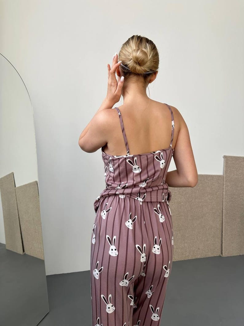 K&ouml;nnte beinhalten: Mauvefarbenes Pyjama-Set mit vertikalem Streifenmuster und wei&szlig;en Hasengesichtern. Das Set besteht aus einem Camisole-Top mit d&uuml;nnen Tr&auml;gern und einer passenden Hose. Die Person ist dem Betrachter abgewandt.