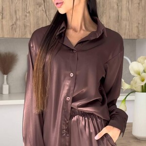 May include: A brown satin pajama set. The long-sleeved button-down shirt and matching pants have a relaxed fit. The model is wearing sunglasses and gold earrings. The background features a wooden wall and a vase of white flowers.