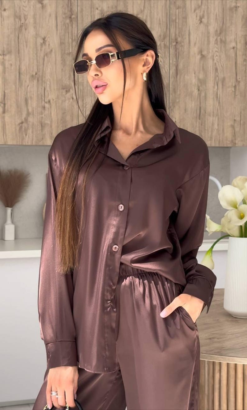 May include: A brown satin pajama set. The long-sleeved button-down shirt and matching pants have a relaxed fit. The model is wearing sunglasses and gold earrings. The background features a wooden wall and a vase of white flowers.