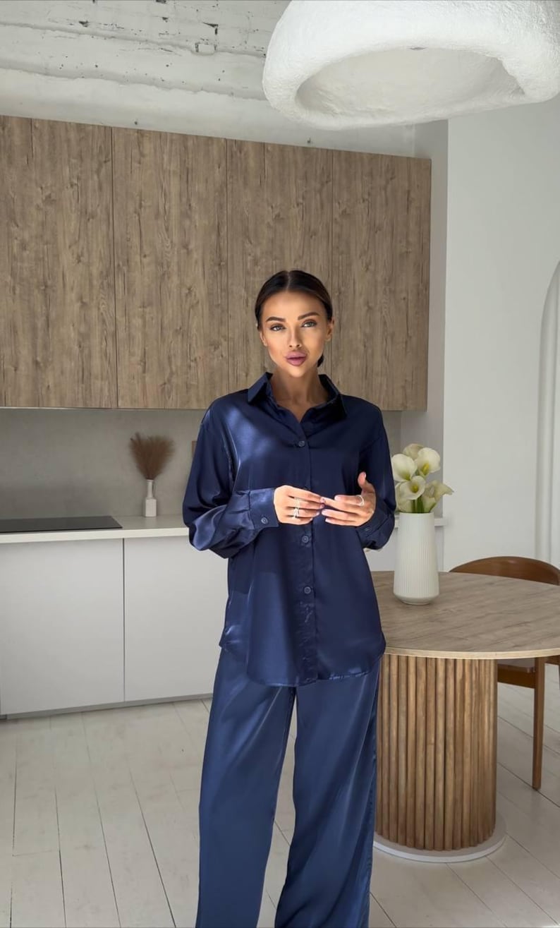 May include: A navy blue satin pajama set. The set includes a button-down shirt and matching wide-leg pants. The outfit is worn in a kitchen setting with a wooden table and white flowers in a vase.