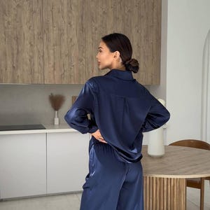 May include: Navy blue satin pajama set. The set includes a long-sleeved button-down shirt and wide-leg pants. The fabric has a subtle sheen. The model is standing in a modern kitchen with light-colored cabinets and a wooden table.