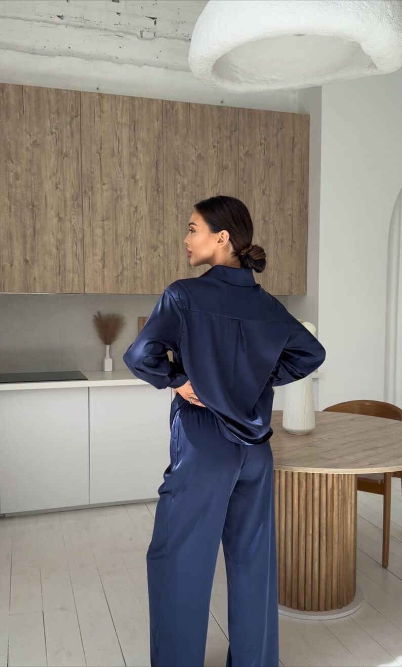 May include: Navy blue satin pajama set. The set includes a long-sleeved button-down shirt and wide-leg pants. The fabric has a subtle sheen. The model is standing in a modern kitchen with light-colored cabinets and a wooden table.