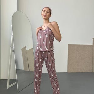 K&ouml;nnte beinhalten: Mauvefarbenes Pyjama-Set mit Hasenmuster, bestehend aus einem Tanktop und einer Hose. Das Set hat vertikale Streifen und wei&szlig;e Hasengesichter. Das Model tr&auml;gt au&szlig;erdem ein passendes Stirnband und schwarze Hausschuhe mit rosa Herzakzenten.