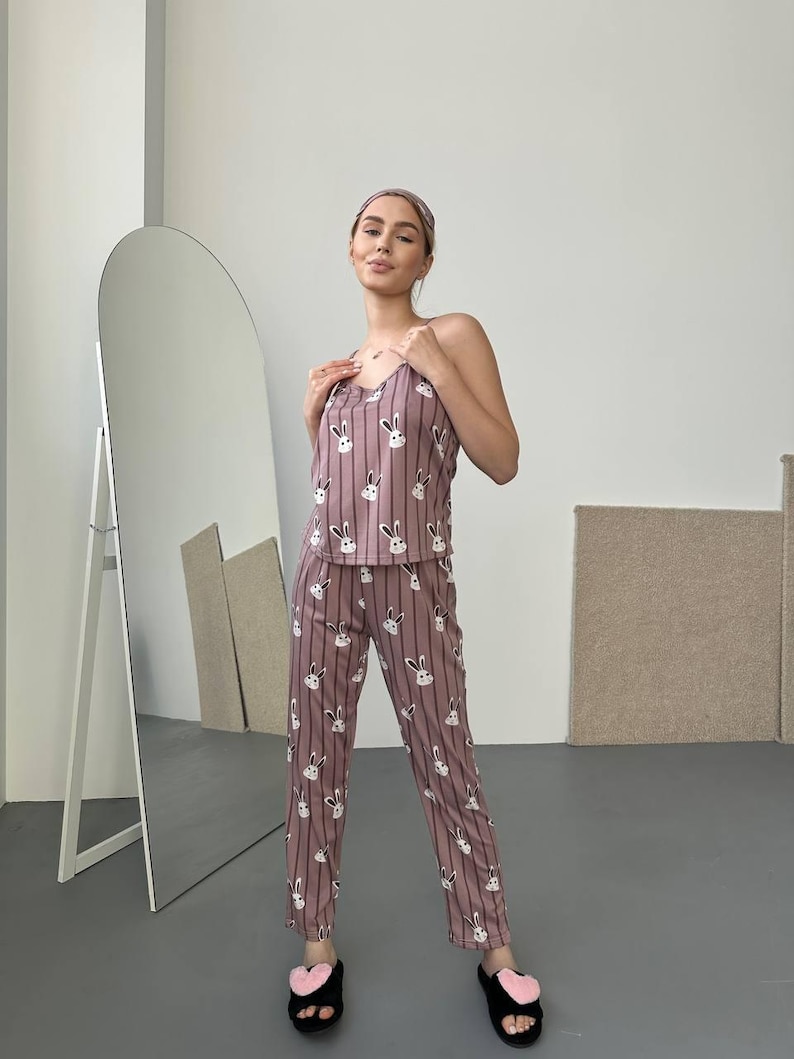 K&ouml;nnte beinhalten: Mauvefarbenes Pyjama-Set mit Hasenmuster, bestehend aus einem Tanktop und einer Hose. Das Set hat vertikale Streifen und wei&szlig;e Hasengesichter. Das Model tr&auml;gt au&szlig;erdem ein passendes Stirnband und schwarze Hausschuhe mit rosa Herzakzenten.