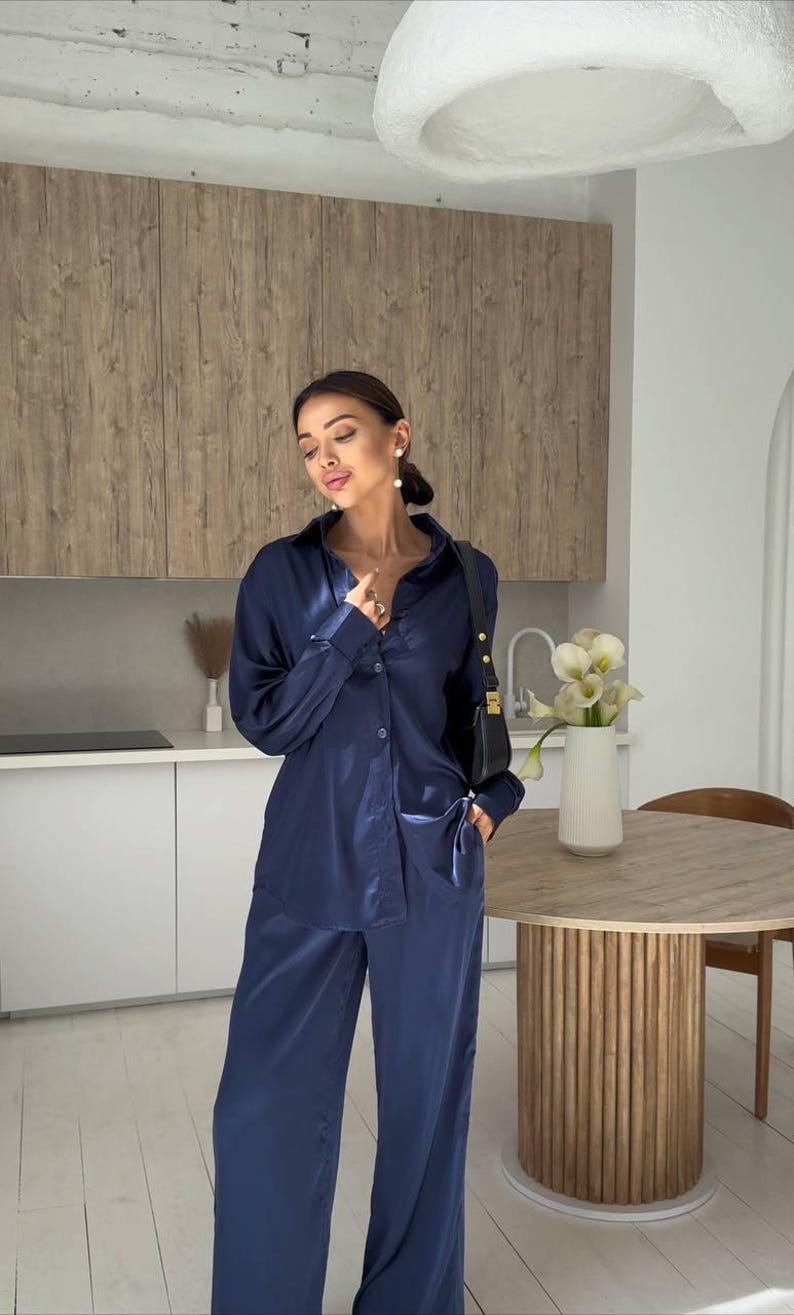 May include: Navy blue satin pajama set. The set includes a button-down shirt with a pocket and wide-leg pants. The outfit is worn with a black shoulder bag. The background includes a kitchen with a wooden table and a white vase with flowers.