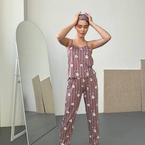 K&ouml;nnte beinhalten: Ein zweiteiliger Schlafanzug in Mauve mit vertikalem Streifenmuster und wei&szlig;en Hasengesichtern. Das Set beinhaltet ein Camisole-Top, eine Hose und ein Stirnband. Die Person tr&auml;gt auch schwarze Hausschuhe mit rosa Herzakzenten.