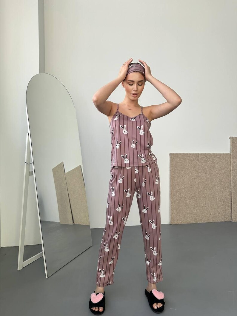 K&ouml;nnte beinhalten: Ein zweiteiliger Schlafanzug in Mauve mit vertikalem Streifenmuster und wei&szlig;en Hasengesichtern. Das Set beinhaltet ein Camisole-Top, eine Hose und ein Stirnband. Die Person tr&auml;gt auch schwarze Hausschuhe mit rosa Herzakzenten.