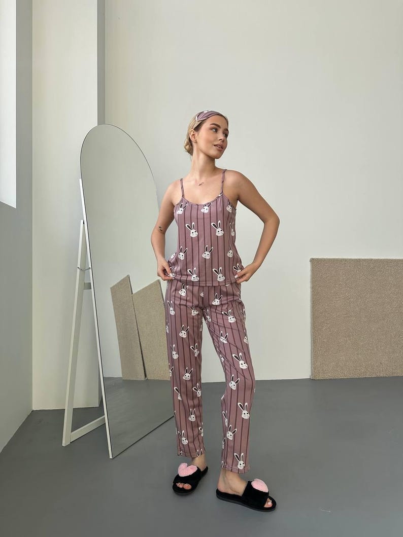 K&ouml;nnte beinhalten: Mauvefarbenes Pyjama-Set mit wei&szlig;en vertikalen Streifen und wei&szlig;en Hasengesichtern. Das Set besteht aus einem Tanktop, einer Hose und einem Stirnband. Die Person tr&auml;gt auch schwarze Hausschuhe mit rosa herzf&ouml;rmigen Details.