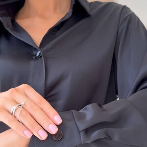 May include: A black button-down shirt with a classic collar and long sleeves. The shirt is made of a smooth, shiny fabric and features buttons on the cuffs and down the front. The overall style is elegant and simple.