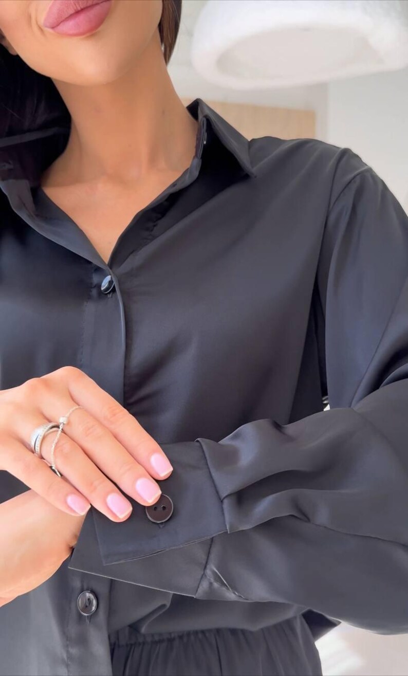 May include: A black button-down shirt with a classic collar and long sleeves. The shirt is made of a smooth, shiny fabric and features buttons on the cuffs and down the front. The overall style is elegant and simple.