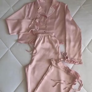 Könnte beinhalten: Ein hellrosa Pyjama-Set, bestehend aus einem geknöpften Oberteil mit Kragen und langen Ärmeln sowie einer passenden Hose mit Kordelzug. Das Set ist aus seidigem Material gefertigt und hat dekorative Rüschen an den Ärmeln und am Hosenbund.