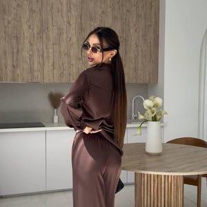 May include: A brown satin pajama set, consisting of a button-down shirt and wide-leg pants. The outfit is worn with sunglasses. The background includes a kitchen with wooden cabinets and a round table.