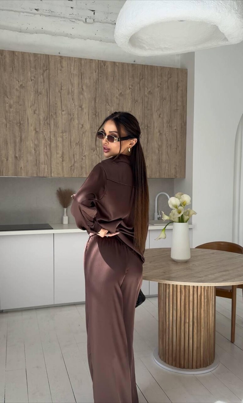 May include: A brown satin pajama set, consisting of a button-down shirt and wide-leg pants. The outfit is worn with sunglasses. The background includes a kitchen with wooden cabinets and a round table.