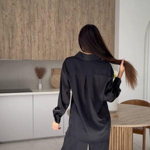 May include: Black satin pajama set. The set includes a long-sleeved button-down shirt and wide-leg pants. The model is holding her hair. The background includes a kitchen with wooden cabinets and a round table.