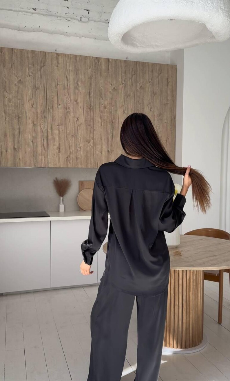 May include: Black satin pajama set. The set includes a long-sleeved button-down shirt and wide-leg pants. The model is holding her hair. The background includes a kitchen with wooden cabinets and a round table.