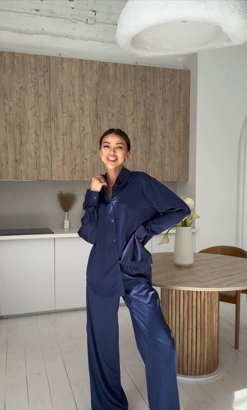 May include: A navy blue satin pajama set, consisting of a button-down shirt and wide-leg pants. The outfit is worn in a kitchen setting with a wooden table and cabinets in the background. The model is smiling.