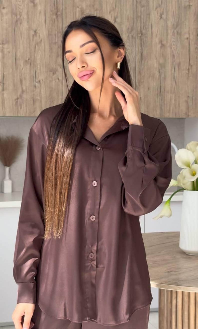 May include: A brown satin button-down shirt with long sleeves. The shirt has a classic collar and is buttoned up. The fabric has a slight sheen. The shirt is part of a matching set.