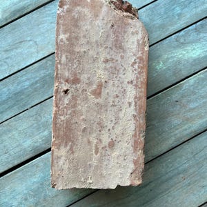 May include: A weathered, rectangular brick with a rough, uneven surface. The brick is a reddish-brown color with white patches of dust and dirt.