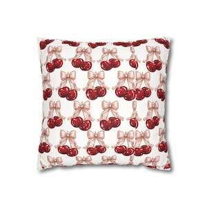 May include: A square throw pillow featuring a repeating pattern of glossy red cherries paired with light beige bows on a white background. The cherries are depicted in a watercolor style with visible highlights, and the bows are tied in a simple knot.