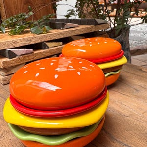May include: Two ceramic burger-shaped containers. The top bun is orange with white sesame seed details. The burger layers include red, yellow, and green sections. The containers sit on a wooden surface.
