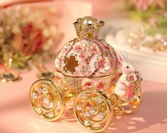 May include: A decorative pink carriage with gold wheels and accents. The carriage is adorned with floral designs and sparkling jewels. A small white rabbit figure is seated on the front, holding a bouquet. A pearl sits atop the carriage.