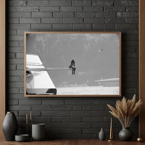May include: Black and white framed photograph of a person sitting on a diving board, with a boat and a bird in the background. The image is set against a backdrop of water and sky, creating a serene and contemplative scene.