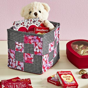 May include: A Valentine's Day gift basket with a white teddy bear, candy, and a heart-shaped box. The basket is black and white checkered with red and pink heart-themed patches. Boxes of Sweethearts candy are visible.