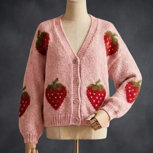 Peut inclure: Un cardigan rose clair avec un col en V et une fermeture boutonnée. Le pull est orné de fraises brodées rouges et vertes sur le devant et les manches. Le vêtement est présenté sur un mannequin.
