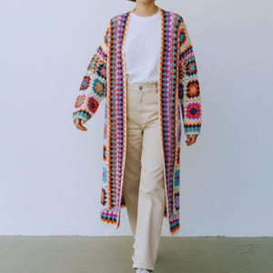 May include: A long, colourful crochet cardigan with a geometric pattern in shades of orange, blue, pink, and white. The open-front sweater is knee-length and features a vibrant, patchwork design, ideal for layering.