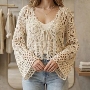 May include: Cream-colored crochet cardigan with long, flared sleeves and a tie closure. The open-weave design features floral and geometric patterns. The cardigan is worn over a white top and paired with blue jeans.