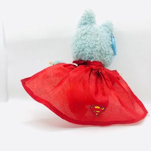Puede incluir: Un juguete de peluche azul claro con forma de gato que lleva una capa roja con el logotipo de Superman. La capa est&aacute; atada alrededor del cuello del gato. El juguete tiene una etiqueta en la oreja.
