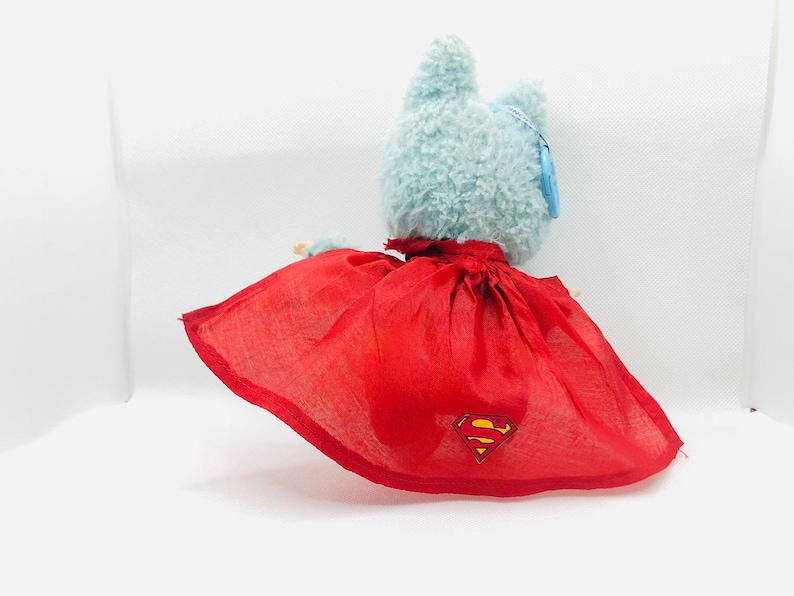 Puede incluir: Un juguete de peluche azul claro con forma de gato que lleva una capa roja con el logotipo de Superman. La capa est&aacute; atada alrededor del cuello del gato. El juguete tiene una etiqueta en la oreja.