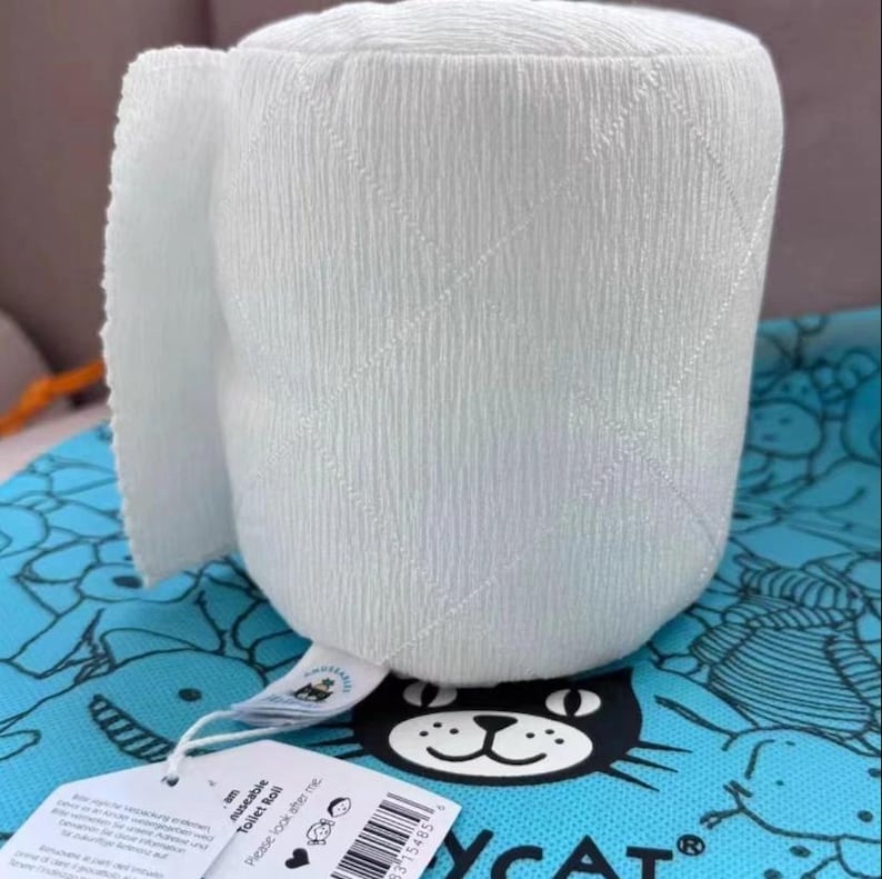 May include: A white, plush toilet roll with a textured surface and a small section of unrolled paper. The roll is sitting on a blue patterned surface with a black cat logo and a tag that reads "reusable Toilet Roll".