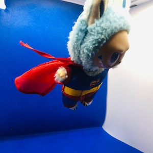 Puede incluir: Un juguete de peluche disfrazado de superh&eacute;roe, con un traje azul y rojo y una capa roja. El juguete tiene una capucha de piel azul claro y una cara de color canela. El fondo es un degradado de azul y blanco.