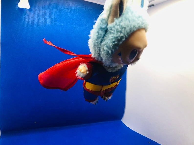Puede incluir: Un juguete de peluche disfrazado de superh&eacute;roe, con un traje azul y rojo y una capa roja. El juguete tiene una capucha de piel azul claro y una cara de color canela. El fondo es un degradado de azul y blanco.