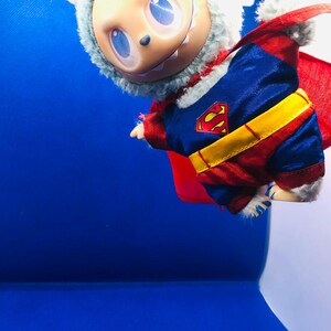 Puede incluir: Una mu&ntilde;eca caprichosa vestida de Superman, con un traje azul y rojo con un cintur&oacute;n amarillo y el logo de Superman. La mu&ntilde;eca tiene una capucha de piel azul claro y una capa roja, sobre un fondo azul.