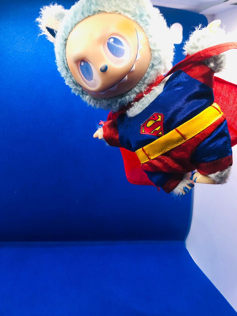Puede incluir: Una mu&ntilde;eca caprichosa vestida de Superman, con un traje azul y rojo con un cintur&oacute;n amarillo y el logo de Superman. La mu&ntilde;eca tiene una capucha de piel azul claro y una capa roja, sobre un fondo azul.