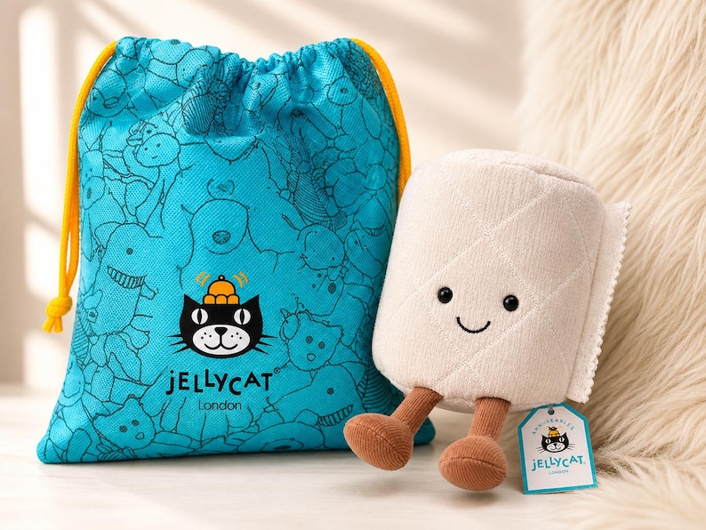 May include: A plush toy roll of toilet paper with a smiling face and brown legs, next to a blue drawstring bag with a pattern of cartoon animals. The bag has a yellow drawstring and the Jellycat London logo.